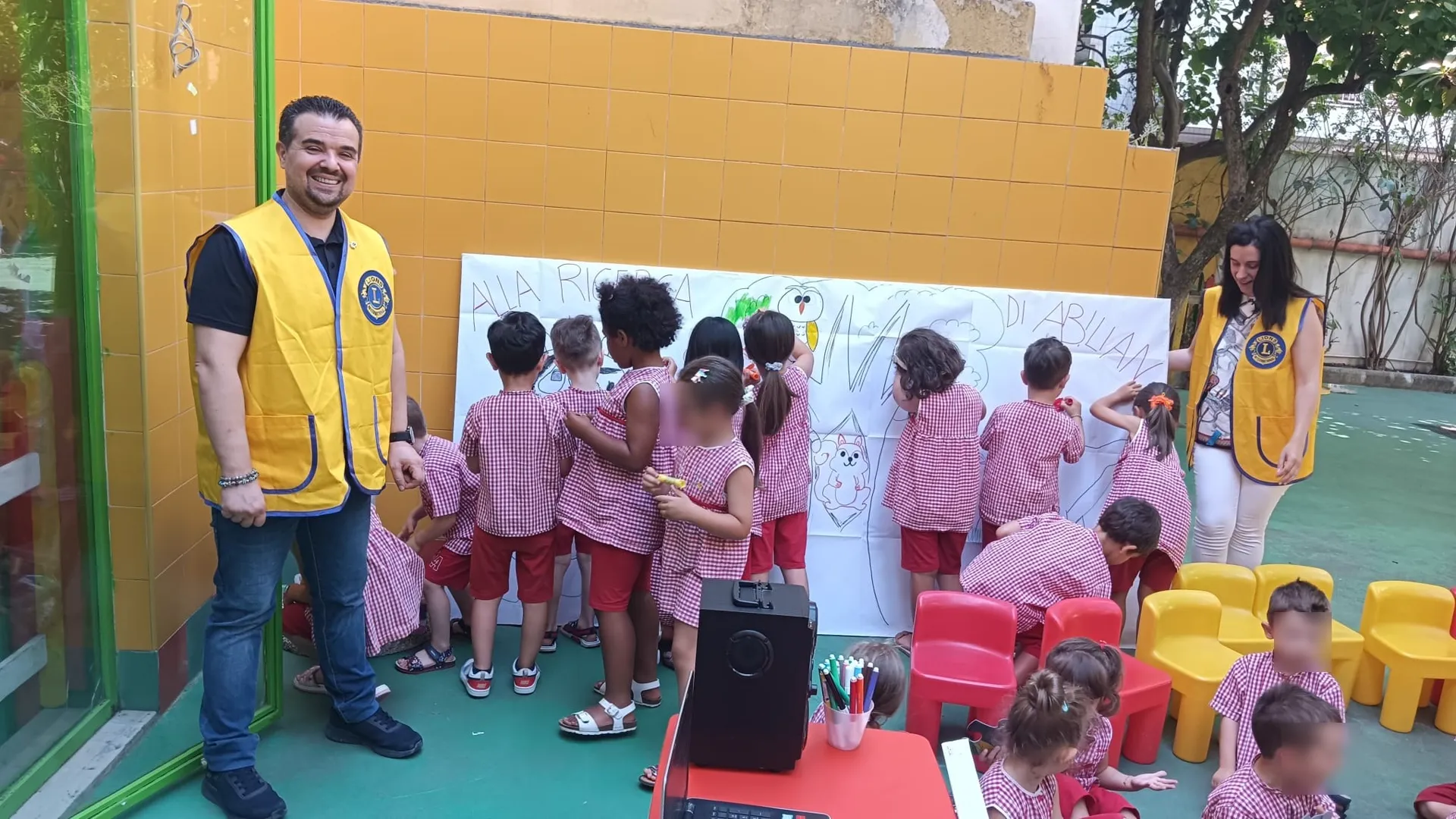 Volontari del Club accanto a un gruppo di bambini che colorano un grande cartellone nel cortile di una scuola dell’infanzia